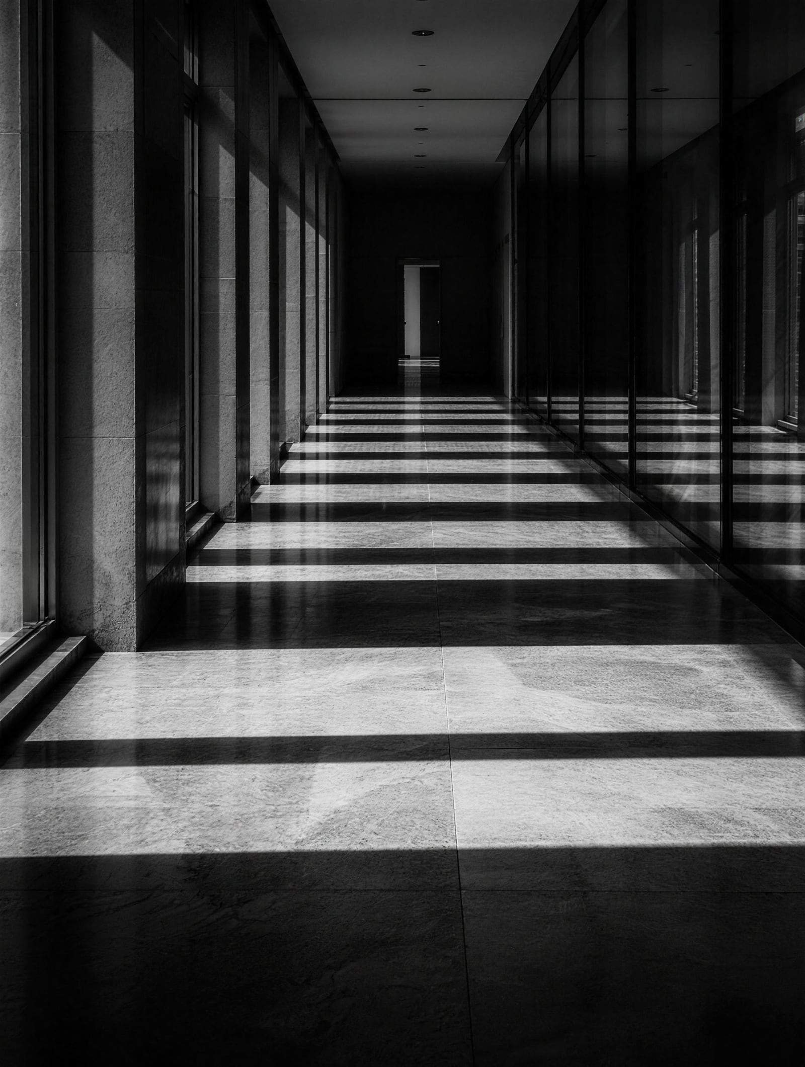 A long corridor of glass and stone, morning light in bars across the floor.