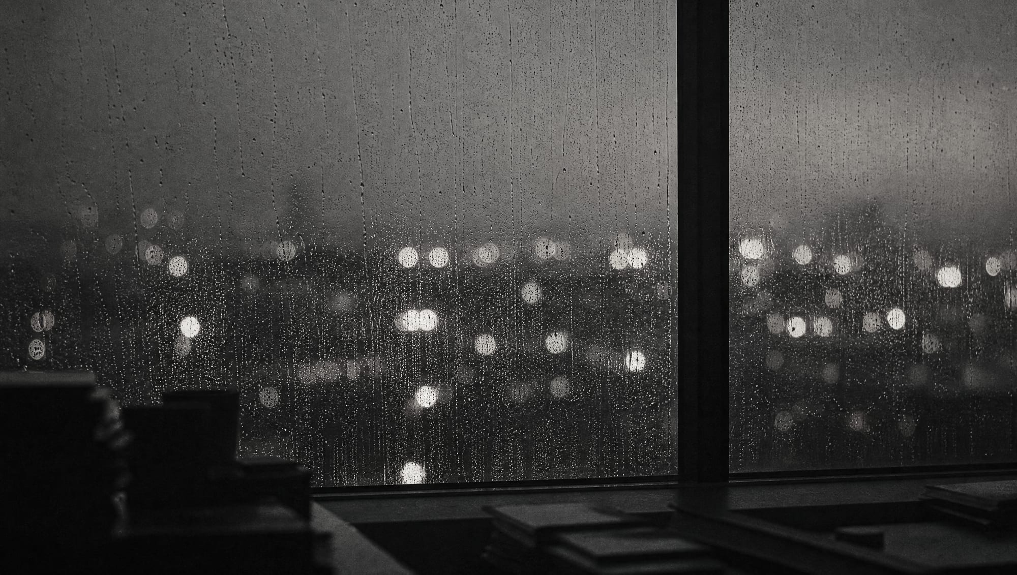 Rain on a large window at dusk seen from a dark office, the lights outside out of focus.